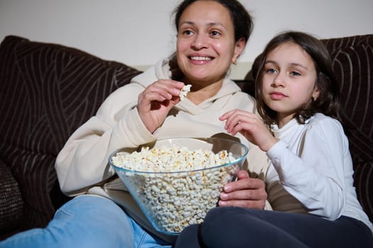 A loving mother and her young daughter are enjoying a bowl of popcorn while seated on a comfortable couch, spending quality time together at home.