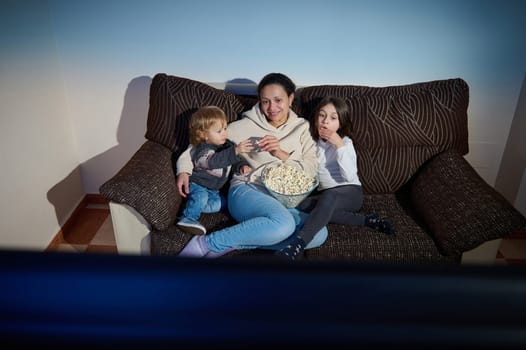 A mother and her two children sit on a couch, sharing popcorn and enjoying entertainment together in a cozy living room setting. Their expressions reflect relaxation, enjoyment, and the warmth of familial bonding.