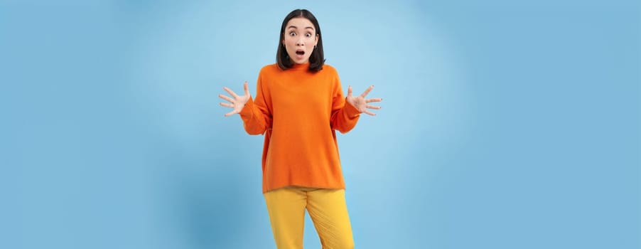 Frustrated young asian woman, shakes hands and looks concerned, panicking, looking worried and upset, standing over blue background.