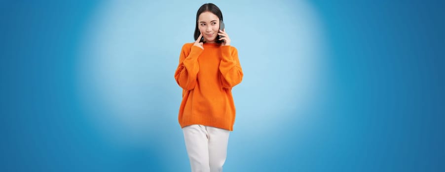 Young woman with thoughtful look, answers phone call, talks on mobile and smiling, thinking with pleased face, standing over blue background.