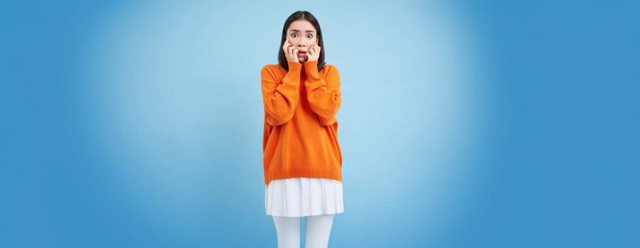 Scared Asian woman shuts her eyes and peeks through hands at smth scary and disturbing, shocked by smth horrifying, standing over blue background.