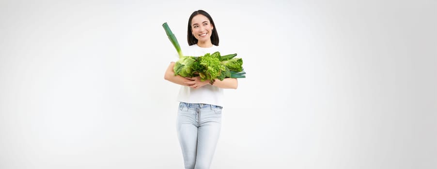 Portrait of happy young woman on green diet, likes eating organic food, holding fresh vegetables from garden, lettuce, cabbage, white studio background.