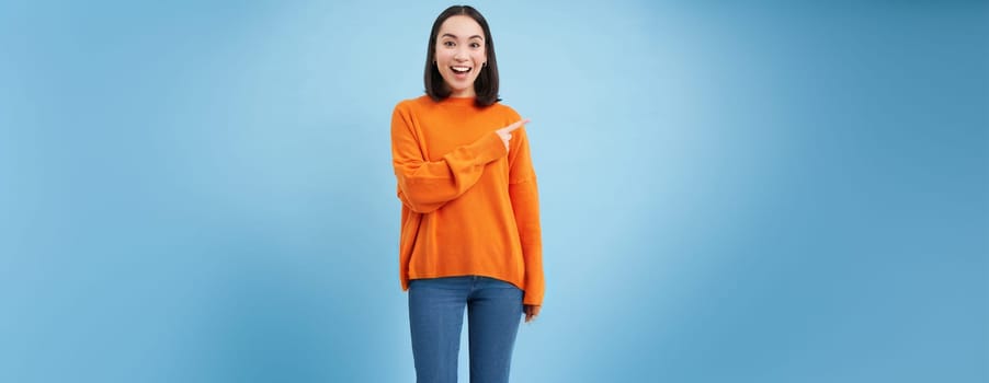 Happy smiling asian woman, pointing finger to the right, shows advertisement, blank copy space on blue background.