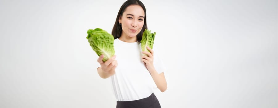 Smiling asian woman, vegan eating cabbage, enjoys tasty fresh lettuce leaves, stands over white background.