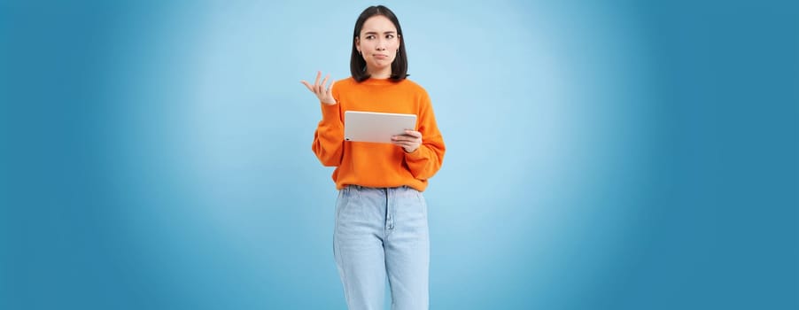 Woman with digital tablet, looks confused, cant understand smth on her gadget, stands puzzled against blue background.