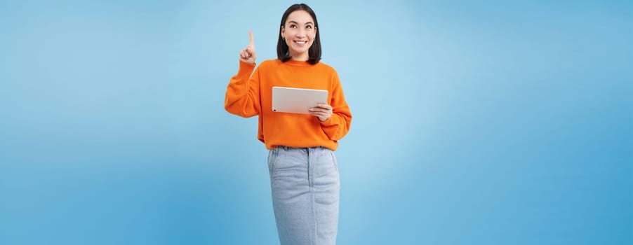 Beautiful young korean woman with tablet, pointing finger up and looking on top advertisement, showing banner, standing over blue background. Online shopping concept.