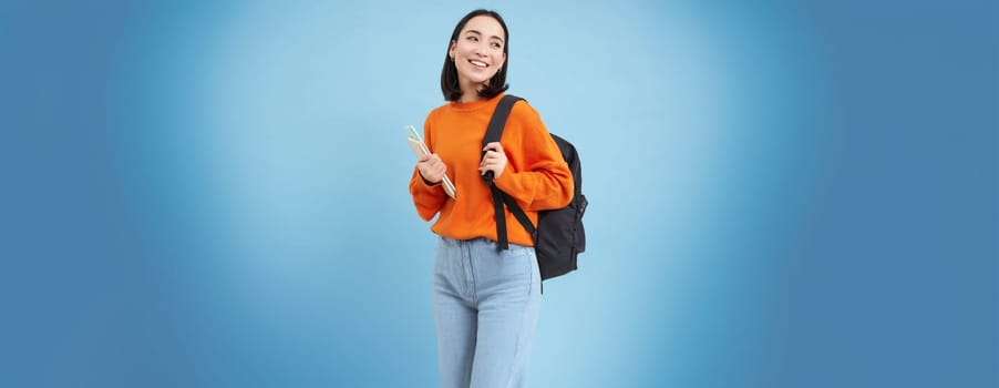 Education and students. Smiling young asian woman with backpack and notebooks, posing against blue background.