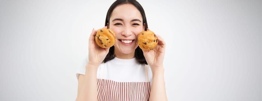 Image of korean woman enjoys eating baked pastry, showing two tasty cupcakes near face and smiles, white background.