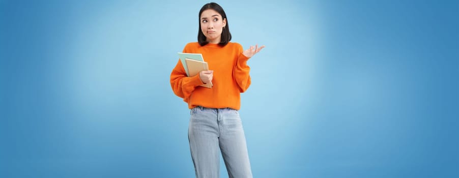 Confused asian woman, student with puzzled face shrugs shoulders, holds notebook, stands over blue background.