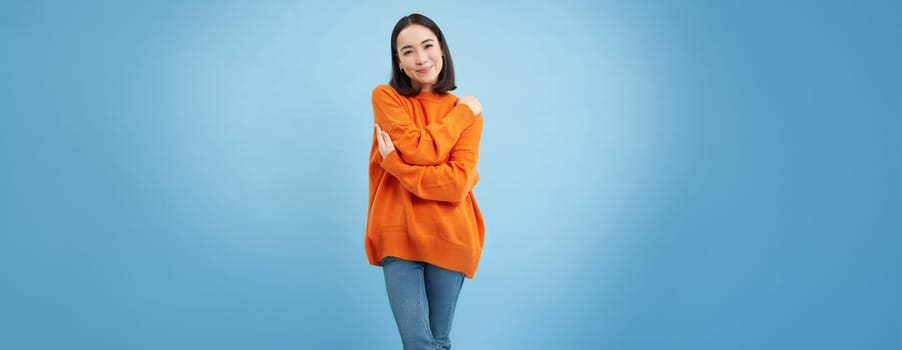 Warmth and tenderness. Cute korean girl hugs herself, embrace equite, femenism and international womens day concept, blue background.