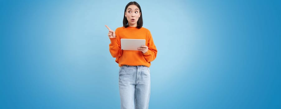 Excited korean woman with digital tablet, pointing at empty space, advertisement banner, holding gadget, showing promo offer, blue background.