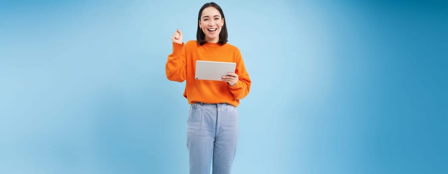 Amazed korean woman with digital tablet, looks surprised and excited, standing over blue background.