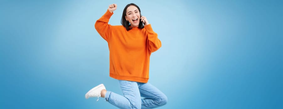 Happy asian girl receives excellent news over the phone, holds mobile and does fist pump, celebrates victory, achievement, blue studio background.