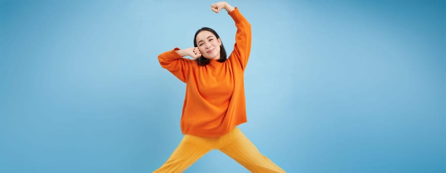 Smiling young korean woman stretching her arms with satisfied face, pleased after good nap, waking up, standing over blue background.