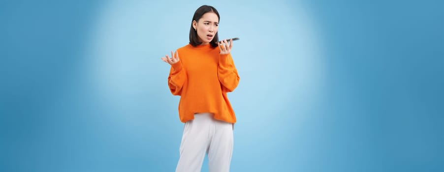 Portrait of upset young woman records voice mail or message, complains with disappointed, sad face, standing over blue background and holding mobile phone.