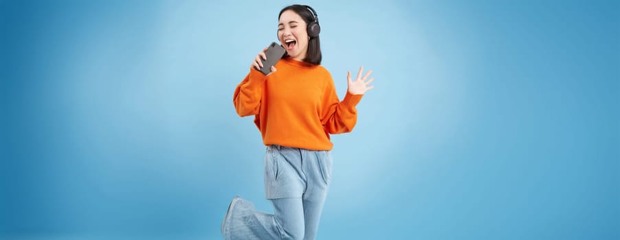 Beautiful korean girl singing in smartphone mic, listens music in headphones, sings karaoke on mobile app, stands over blue background.
