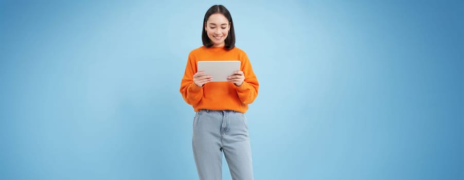 Smiling beautiful asian woman reading on digital tablet, watching videos on gadget or playing video games, standing over blue background.