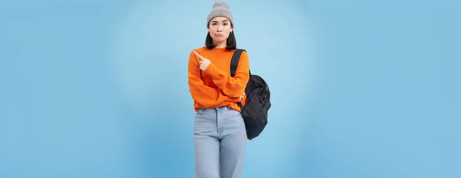 Skeptical asian woman pointing left at strange banner, looks doubtful, stands over studio background.