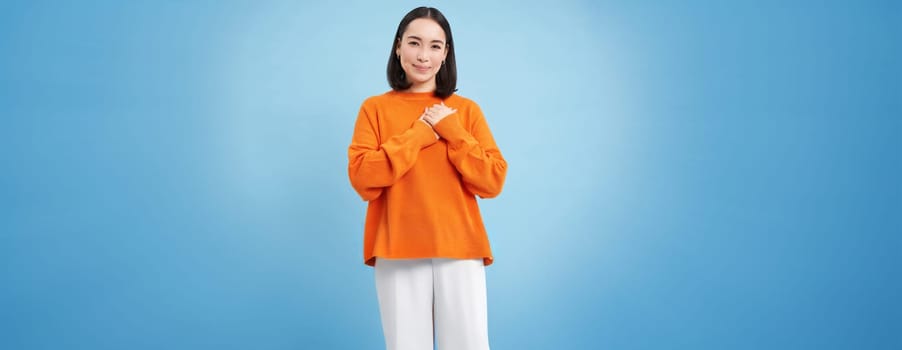 Care and love. Asian woman holds hands on heart and smiles, looks with tenderness and warmth at camera, stands over blue background.