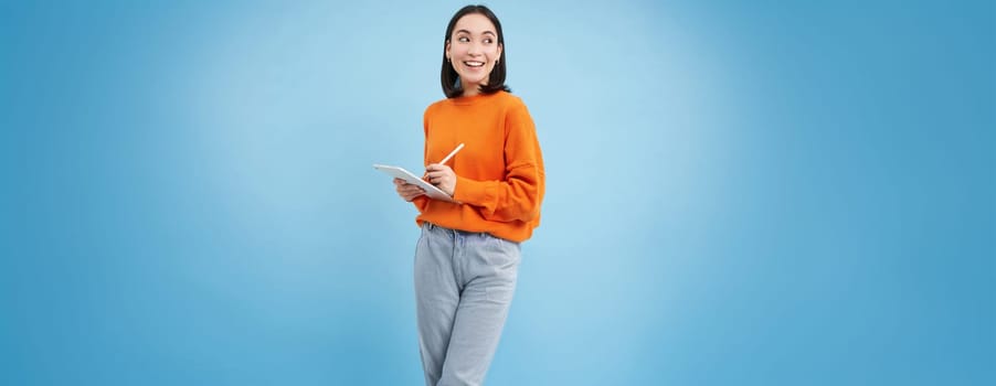 Portrait of young Chinese woman, teacher or student with digital tablet and pencil, writing, taking notes, doing her homework, standing over blue background.
