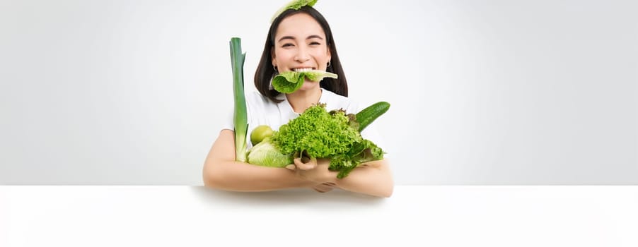 Portrait of smiling korean woman eats lettuce leaf, holding green vegetables, eating nutrisious food, white background.