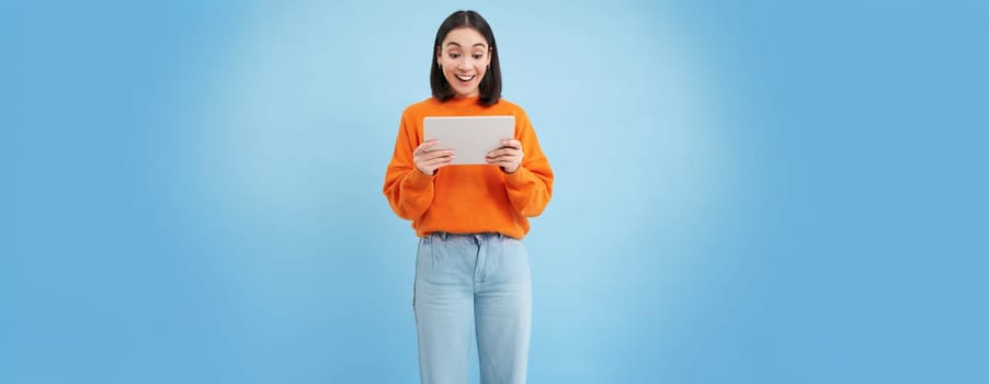 Smiling beautiful asian woman reading on digital tablet, watching videos on gadget or playing video games, standing over blue background.