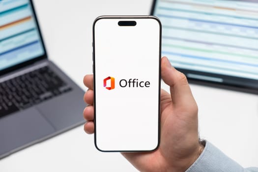 PRAGUE, CZECHIA - JULY, 2024: Microsoft Office logo on the screen of smartphone in mans hand and laptop on the background