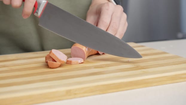 Close-up of a woman cutting sausage on a wooden cutting board in a kitchen setting with different ingredients