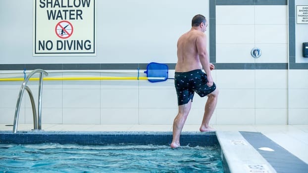 A man in black shorts is getting out of the blue swimming pool. The pool is surrounded by white tiles and there is a sign on the wall saying shallow water no diving.