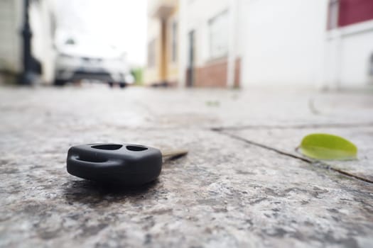 A lost car key rests on the pavement near buildings and foliage.