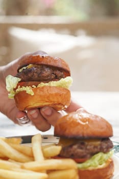 A person holds a burger while fries are placed nearby on a table.