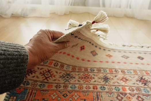 A person rolls up a vibrant patterned rug indoors with natural light.