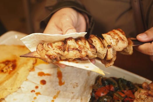 Person enjoys a grilled chicken skewer on a tortilla in a cozy setting.