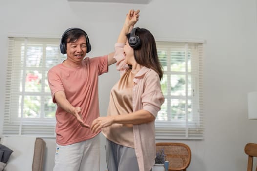 A happy older couple dances together in their living room, wearing headphones and enjoying the music while sharing a joyful moment.