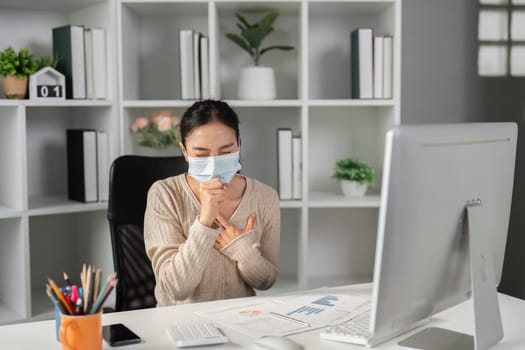A woman wearing a mask coughing while seated at her office desk, reviewing financial reports on her computer.