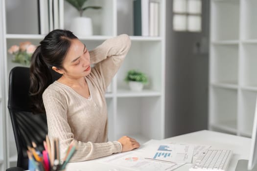 A woman in a modern office experiencing neck discomfort while examining financial reports at her desk.