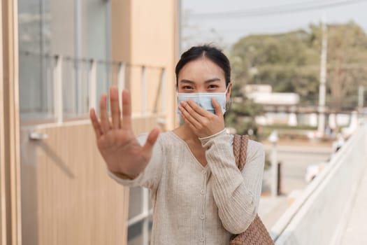 A diverse woman wearing a mask, smiling and signaling stop with her hand outdoors in a city environment.