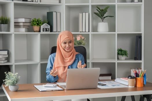 A joyful Muslim woman in a hijab engaging in a video call while working on her laptop at a well-organized office desk.