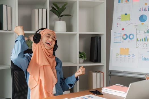 A diverse Muslim woman in a hijab wearing headphones and joyfully celebrating at her desk in a modern office, surrounded by charts and reports.