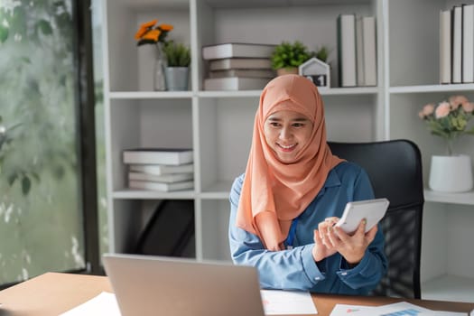 A Muslim woman in a hijab analyzing financial data on her laptop and using a calculator in a contemporary office space.