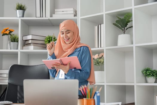A diverse female accountant wearing a hijab joyfully celebrates a financial breakthrough while reviewing her notes in a modern office.