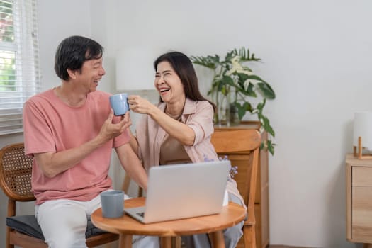 A happy older couple shares a joyful toast with their coffee mugs while enjoying online activities together in their comfortable home.