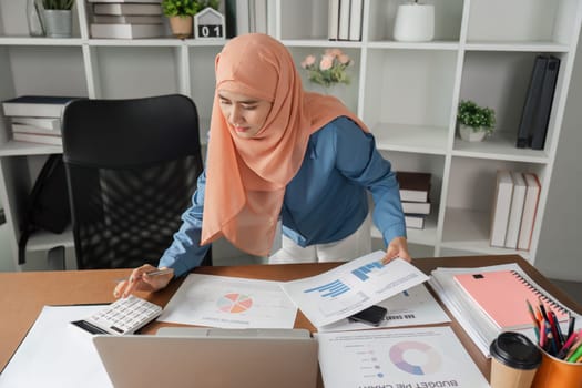A diverse female accountant wearing a hijab reviewing budget reports and calculating finances in a modern office environment.