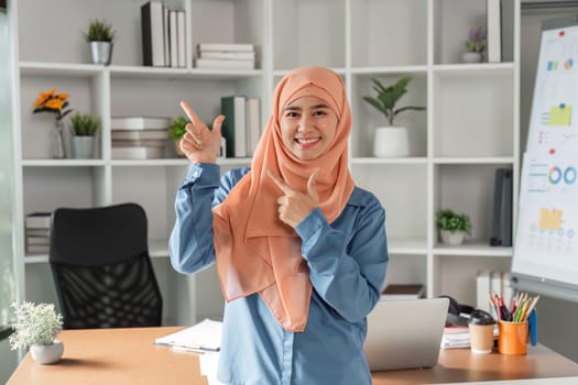 A Muslim woman in a hijab is joyfully pointing with both hands while smiling in her modern office, surrounded by plants and business materials.