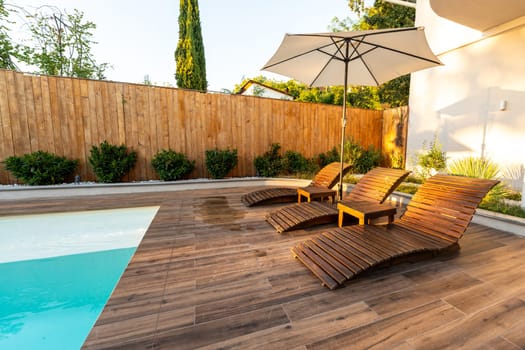 Modern wooden poolside deck with curved lounge chairs, umbrella, and turquoise pool in a sunny backyard
