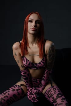 A woman with red hair poses confidently in pink lingerie, expressing allure in a dimly lit setting.