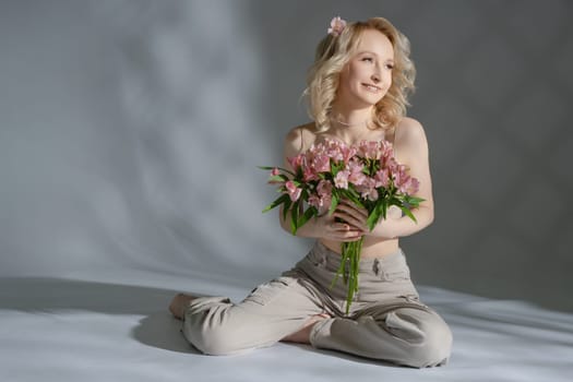 A woman with light blonde hair holds a bouquet of pink flowers, smiling while seated on a smooth surface in gentle lighting.