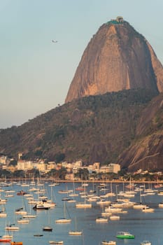 This is a vertical photo of Rio's Sugarloaf Mountain, with a bay full of boats and a plane departing from Santos Dumont airport under a clear blue sky.