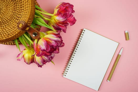 On a pink background there is a notebook, pen, glasses and tulips in a woven straw bag. High quality photo