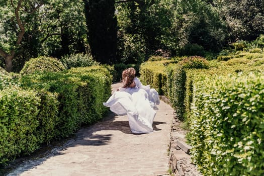 Brunette runs white dress park. A beautiful woman with long brown hair and a long white dress runs along the path along the beautiful bushes in the park, rear view.